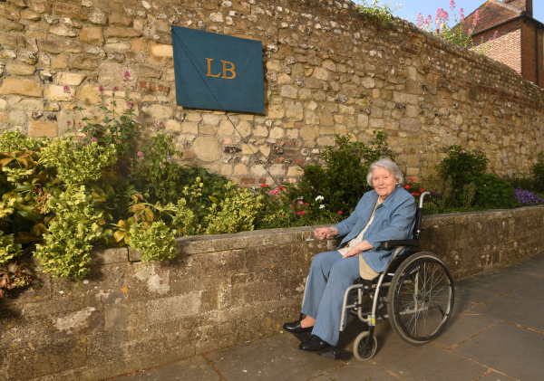This photo shows an elderly woman, Dame Patricia Routledge, seated in a wheelchair along a charming stone pathway beside a historical stone wall adorned with plants and flowers. A cloth cover marked with the initials LB is hanging on the wall, concealing a commemorative plaque.