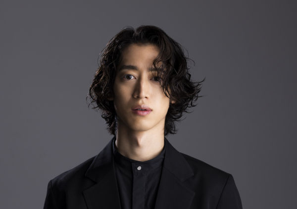 The image features a young Japanese man with shoulder-length wavy black hair, wearing a stylish black suit with a black button-up shirt underneath. He has a calm and composed expression, with his lips slightly parted and his gaze directed at the camera. The background is a neutral gray.