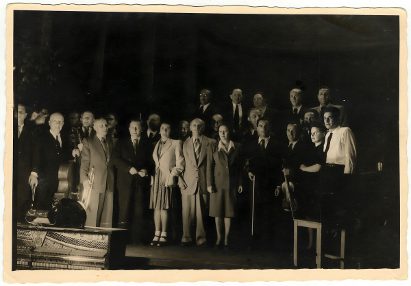 This is a black-and-white photograph depicting a group of musicians, posed together on a stage. The group includes men and women dressed in formal and semi-formal attire, some holding instruments, such as a cello, violin, and a trumpet. A piano is also prominently visible in the foreground. 