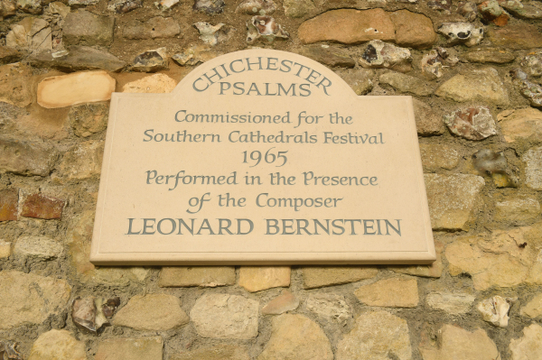 The photo shows a commemorative stone plaque mounted on a stone wall. The inscription on the plaque reads Chichester Psalms, Commissioned for the Southern Cathedrals Festival, 1965, Performed in the Presence of the Composer, Leonard Bernstein